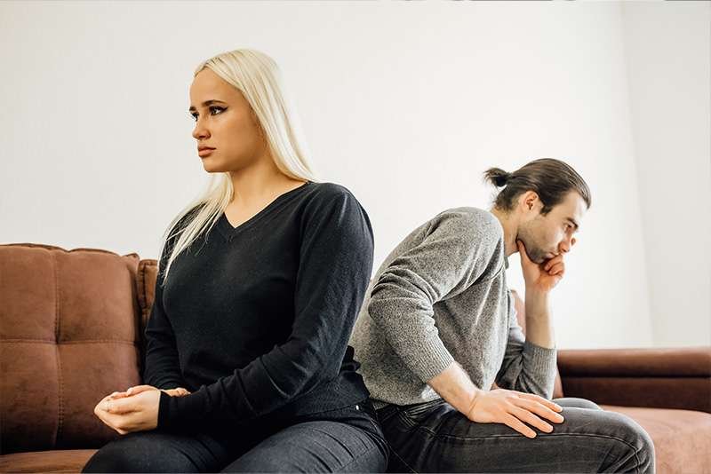 Couple sitting apart on couch, looking upset.